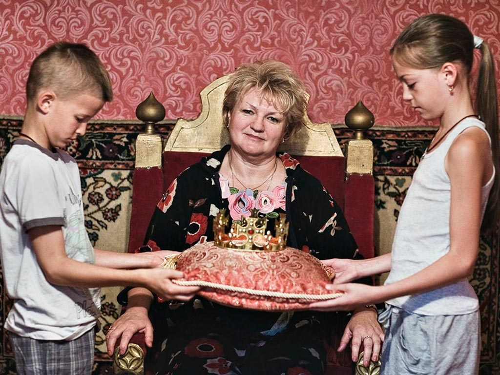 Постановочный снимок: женщина сидит в кресле-троне, мальчик и девочка подносят ей корону на подушке